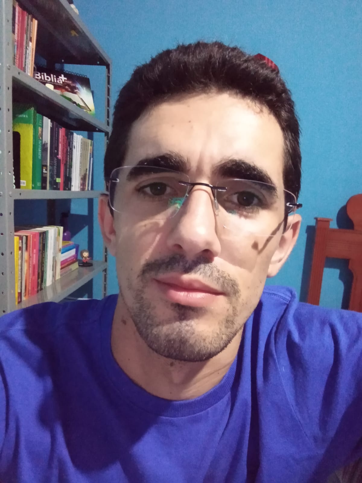 estou usando um óculos de lente solta. camiseta azul sem estampa. Sou branco de cabelos negros, atrás de mim há uma prateleira de livros.