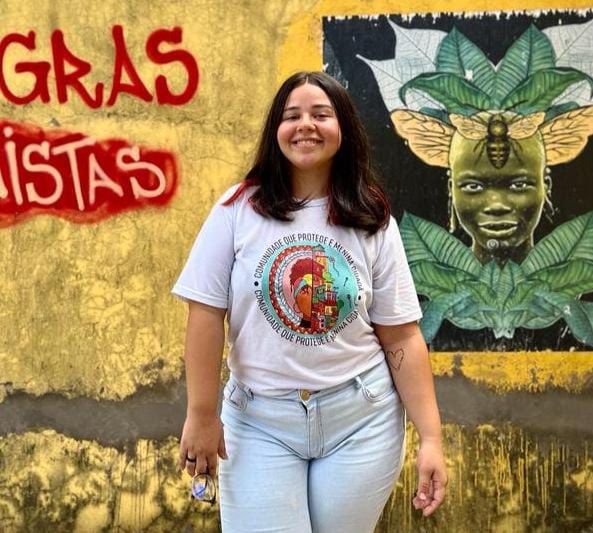 A imagem mostra uma jovem mulher sorridente com cabelos longos e escuros, vestindo uma camiseta branca com uma estampa colorida no centro, e calças jeans claras. Ela está em pé em frente a um muro amarelo com grafites. À esquerda, em vermelho, está escrito "NEGRAS ATIVISTAS". À direita, há uma arte de rua que retrata um rosto humano com asas de abelha e folhas ao fundo.