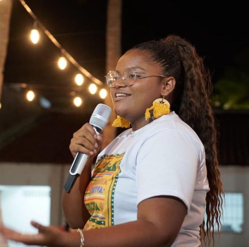 A imagem mostra uma jovem mulher negra sorridente segurando um microfone. Ela está vestindo uma camiseta branca com uma estampa colorida no centro e usa brincos grandes e amarelos com franjas. Ela também usa óculos e tem os cabelos longos presos em um rabo de cavalo. Ao fundo, há luzes decorativas acesas, sugerindo que a foto foi tirada à noite, possivelmente em um evento ao ar livre.