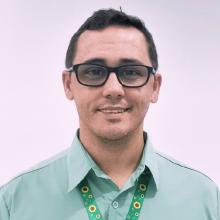 Fernando Costa, homem cisgênero, de pele branca, cabelos e olhos castanhos, deficiente visual. Utiliza óculos de grau, está vestindo a camisa verde Vale e o colar de girassol. Ao fundo uma parede branca.